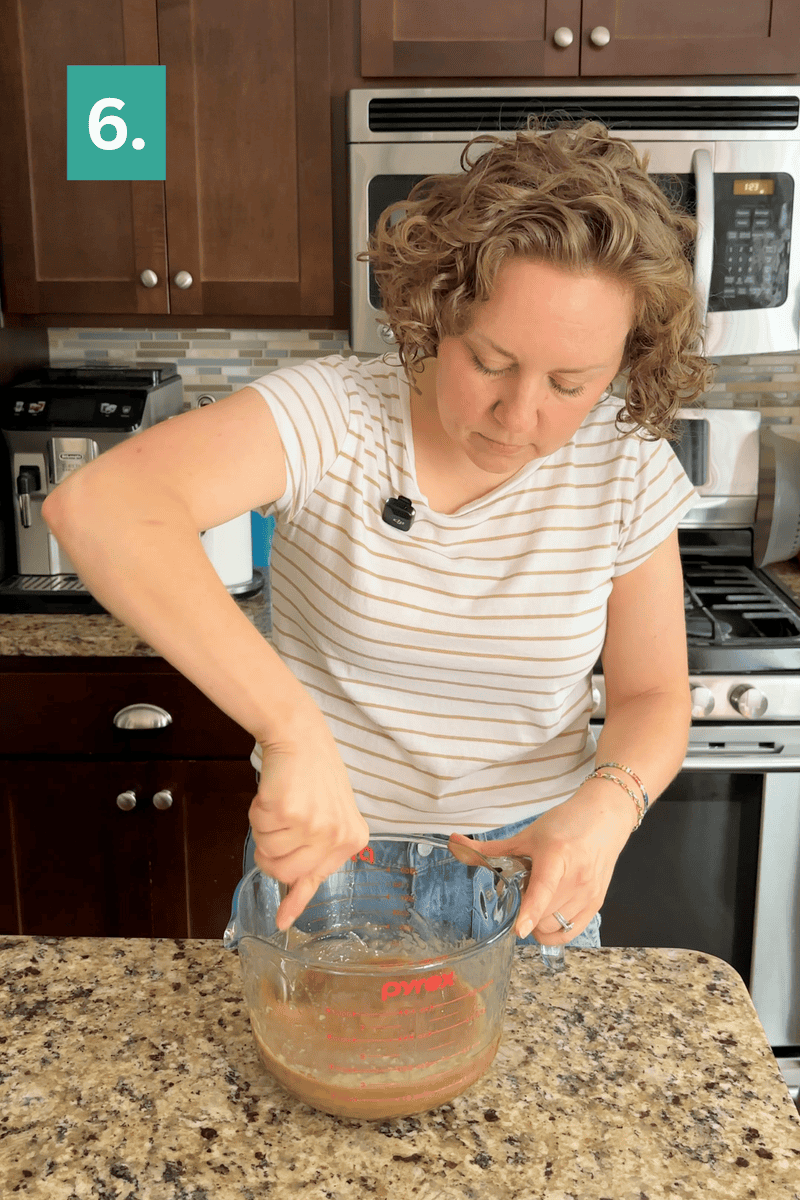 A woman in a striped shirt stirs liquid in a glass measuring cup on a granite kitchen counter. A green square with the number 6 is in the top left corner. Kitchen appliances and cabinets are in the background.