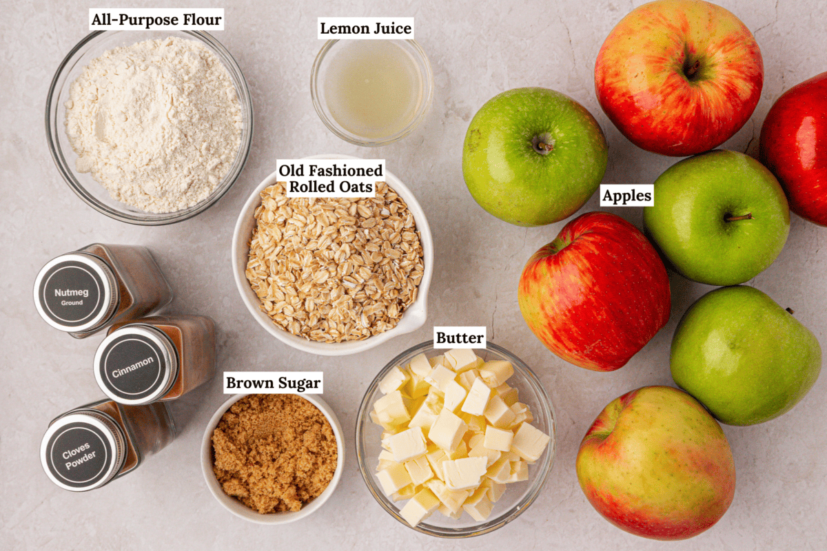 Top-down view of ingredients for apple crisp: apples, diced butter, brown sugar, old fashioned rolled oats, all-purpose flour, lemon juice, and small jars of ground nutmeg, cinnamon, and cloves, all labeled.