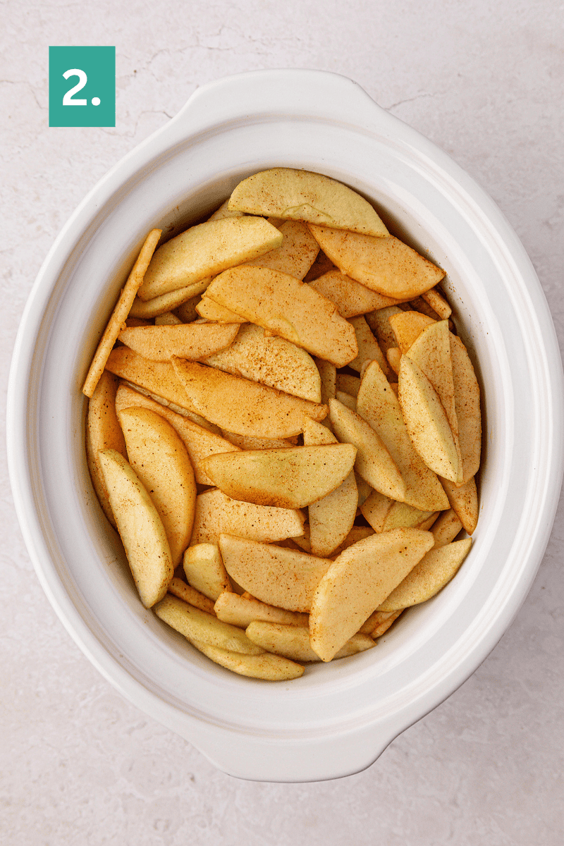 Sliced apples coated with cinnamon and sugar are arranged in a white slow cooker on a light countertop. A green box with the number 2 is in the top left corner.
