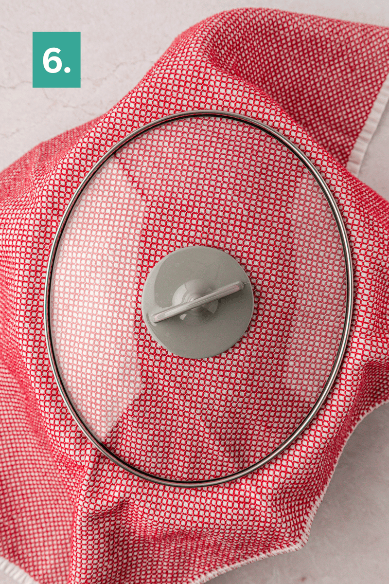 A pot covered with a red patterned cloth and a glass lid sits on a light surface. A green square with the number 6 appears in the upper left corner.