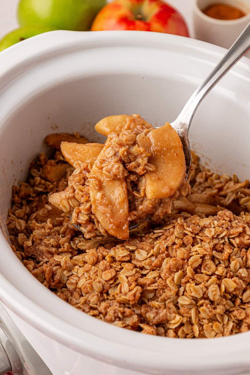 A spoon scoops out apple crisp with oat topping from a white slow cooker. Fresh apples and a small bowl of cinnamon are visible in the background.