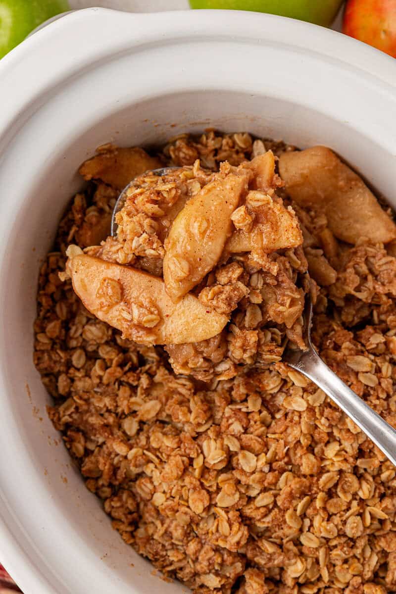 A spoon scoops up baked apple crisp with oat topping from a white dish. Slices of tender, cinnamon-coated apples are visible beneath the golden, crumbly oat topping.