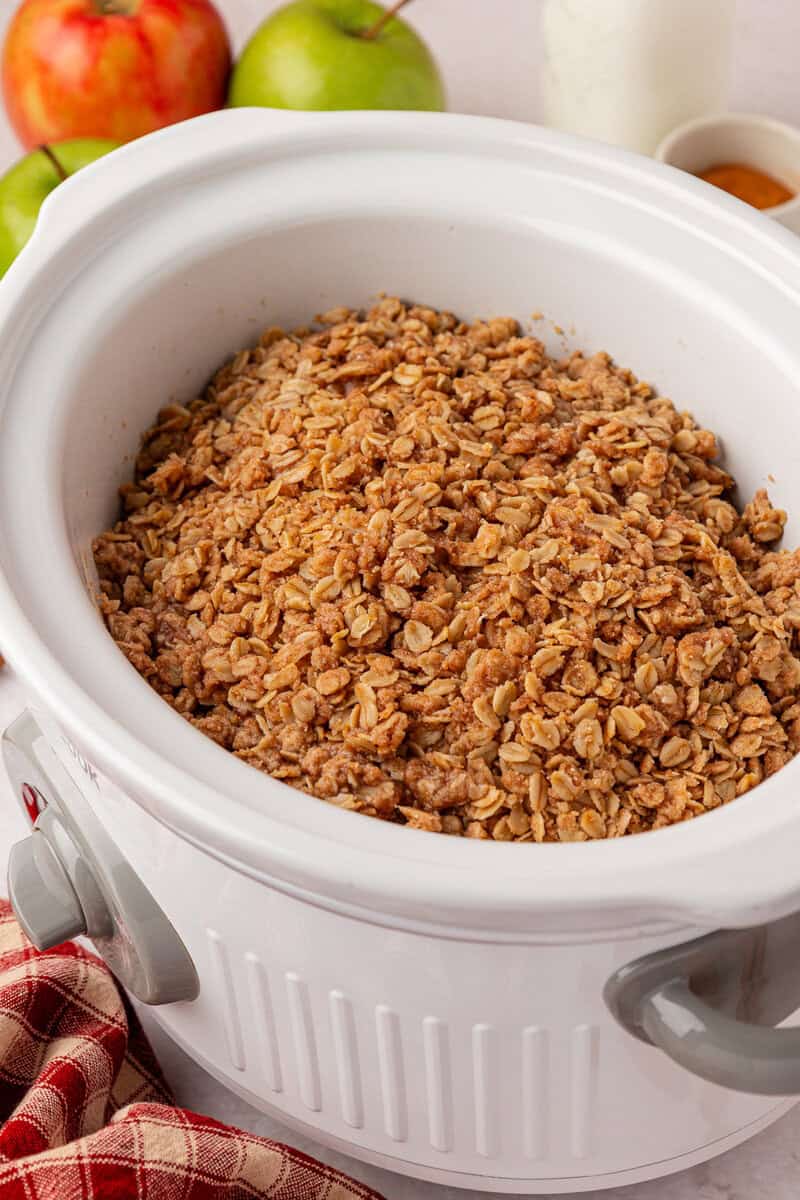 A white slow cooker filled with oat and cinnamon apple crisp, surrounded by green apples, a red apple, and a red plaid towel.