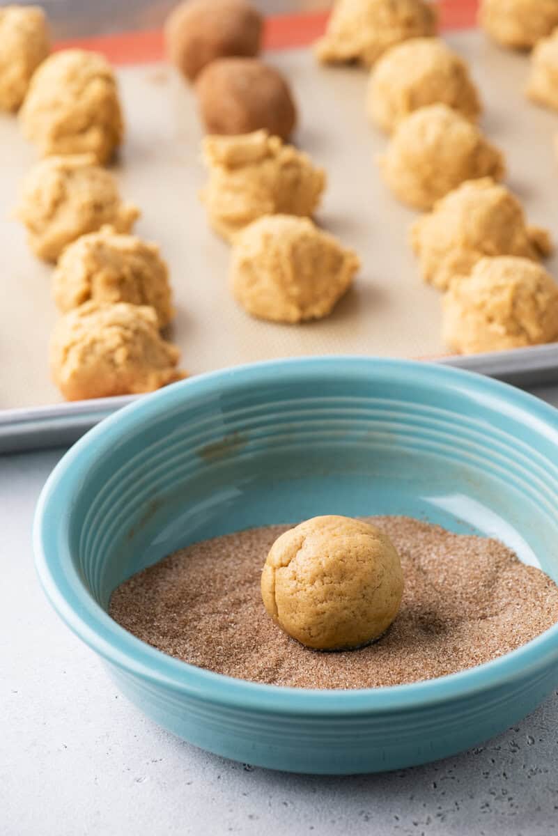 A ball of cookie dough sits in a blue bowl filled with cinnamon sugar, with more dough balls on a baking sheet in the background.