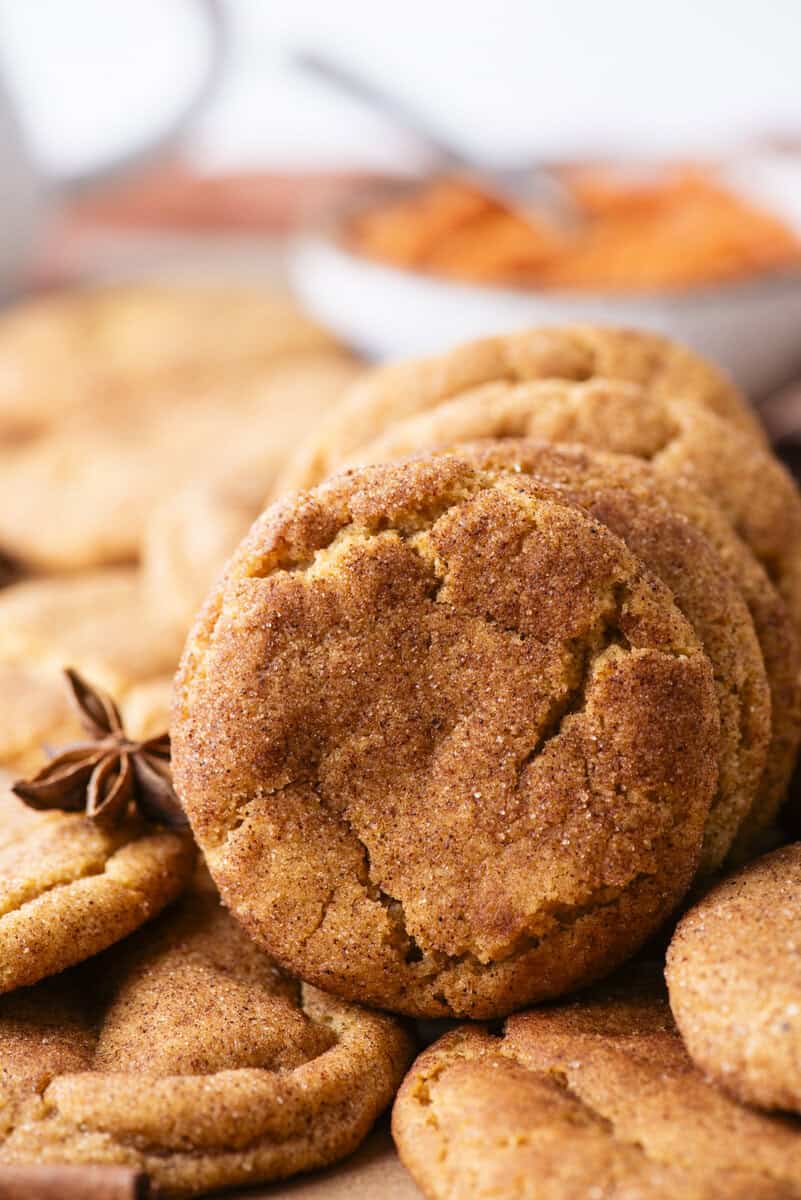 A close-up of several golden brown snickerdoodle cookies, with one cookie standing upright in front, and a blurry bowl of orange filling and a star anise in the background.