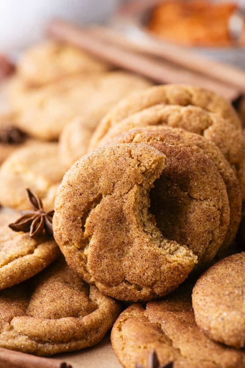 A close-up of snickerdoodle cookies stacked on each other, with one cookie showing a bite taken out. Star anise and cinnamon sticks are scattered around.