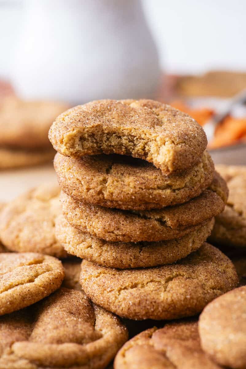 A stack of cinnamon sugar cookies, with the top cookie showing a bite taken out. More cookies are scattered around on a wooden surface.