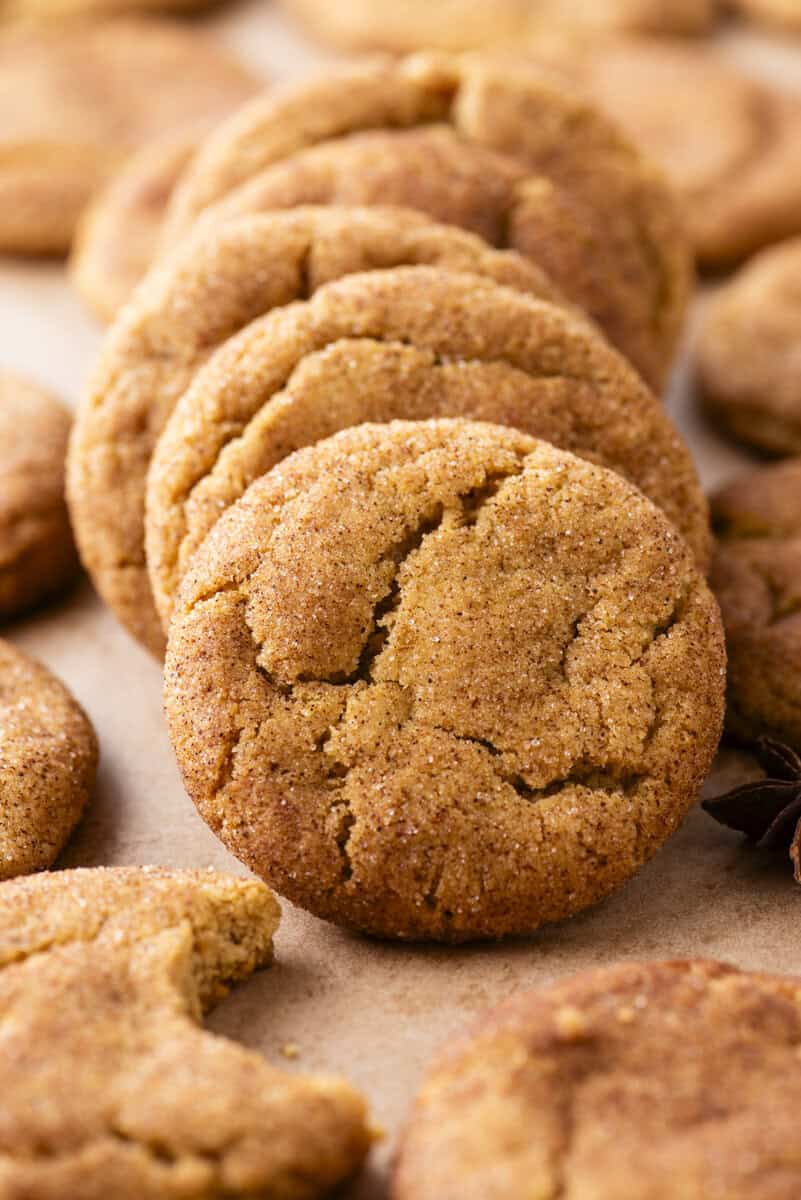 A close-up of several golden-brown, sugar-coated cookies stacked and scattered on a light surface. One cookie in front displays a cracked top, with another partially eaten cookie nearby.