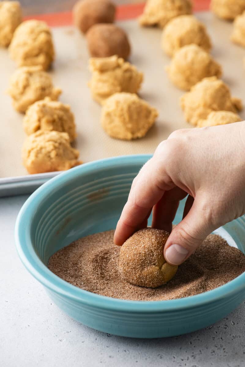 A hand rolls a dough ball in cinnamon sugar in a blue bowl, with more dough balls on a baking sheet in the background.