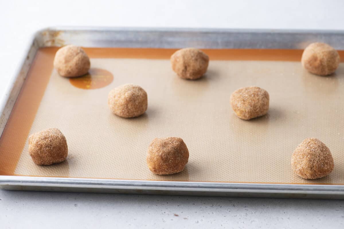 Eight balls of cookie dough coated in cinnamon sugar are evenly spaced on a baking sheet lined with a silicone mat, ready to be baked.