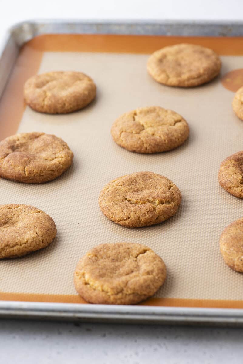 A baking sheet with nine evenly spaced round cookies on a tan silicone baking mat, with a light background. The cookies appear golden brown and slightly cracked on top.