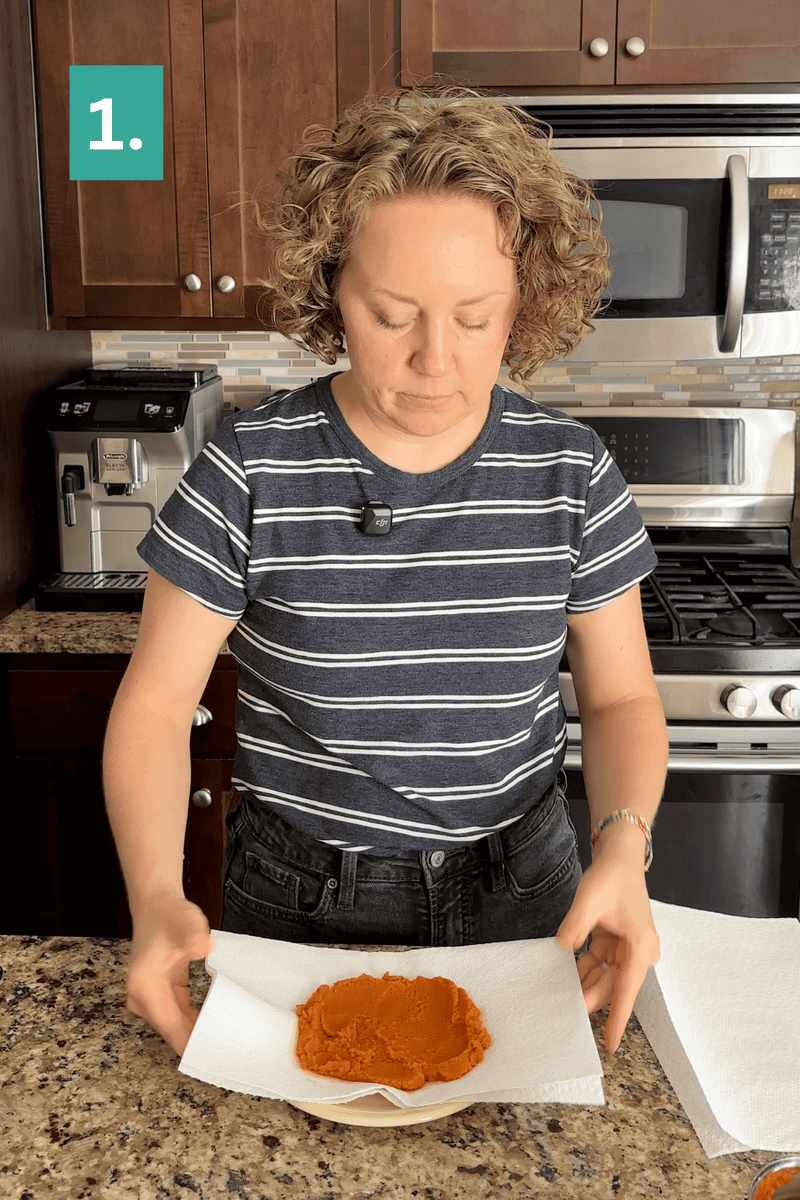 A person in a striped shirt presses pumpkin puree on a plate with paper towels in a kitchen. A green square with the number 1. appears in the top left corner.