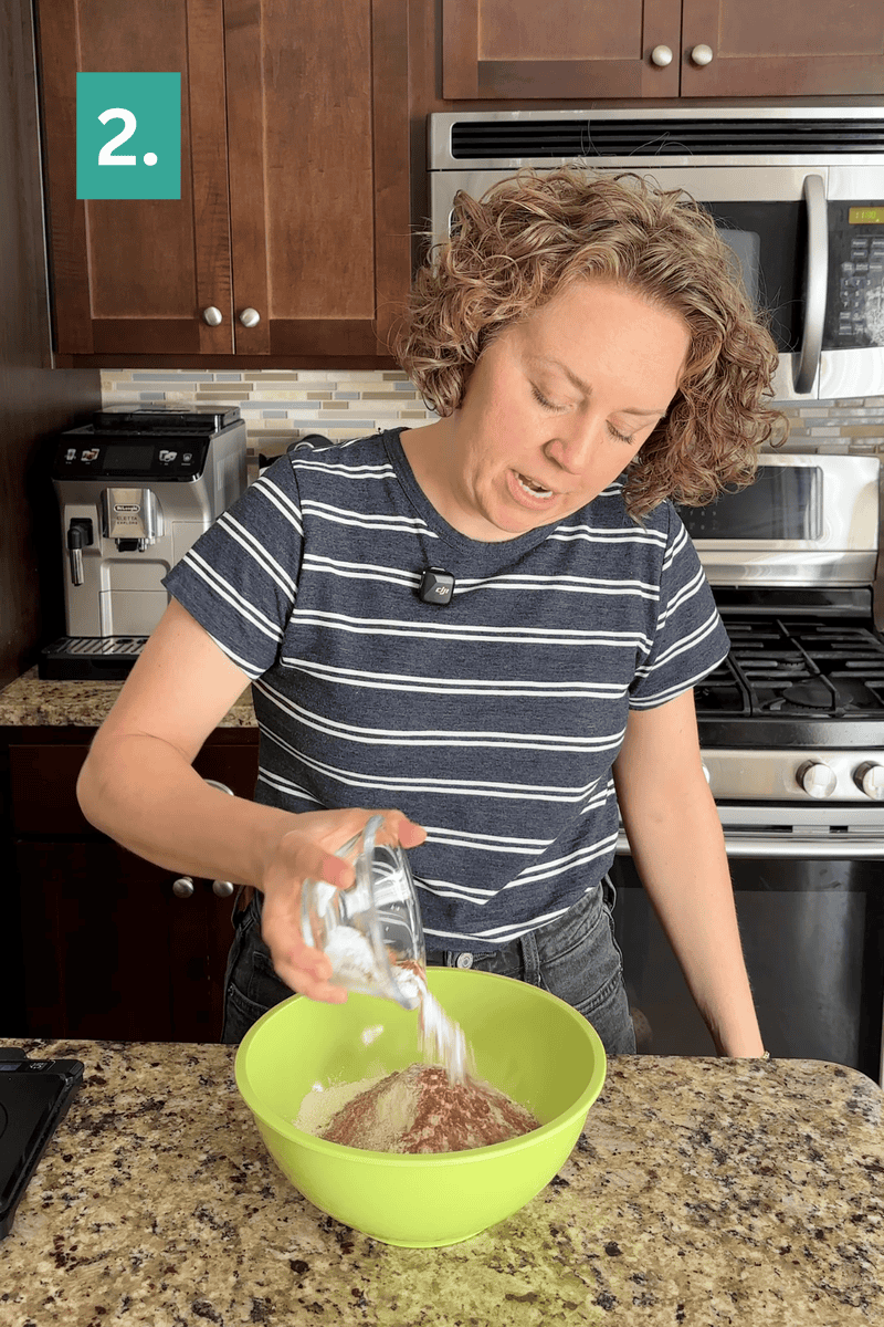A person in a striped shirt pours a cup of white powder into a green mixing bowl with dry ingredients on a kitchen counter. A green box with 2. is in the top left corner.