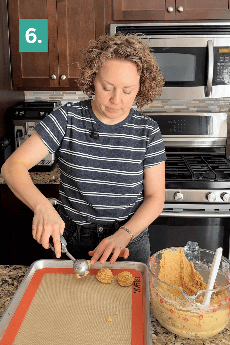A person scoops cookie dough onto a baking tray in a kitchen. A bowl of dough and a spatula are nearby. A green box with the number 6 appears in the top left corner.