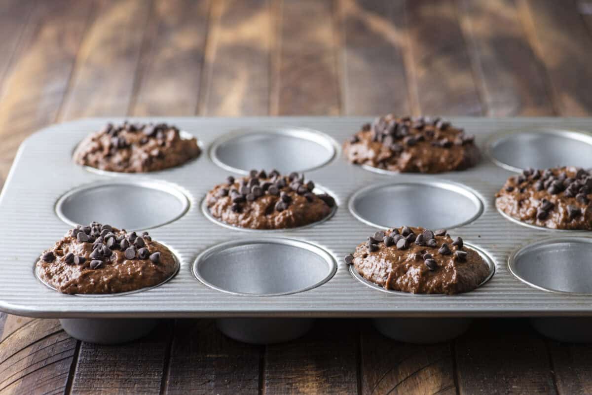 A muffin tin on a wooden surface holds six chocolate muffin batters topped with chocolate chips, while the other six sections are empty.