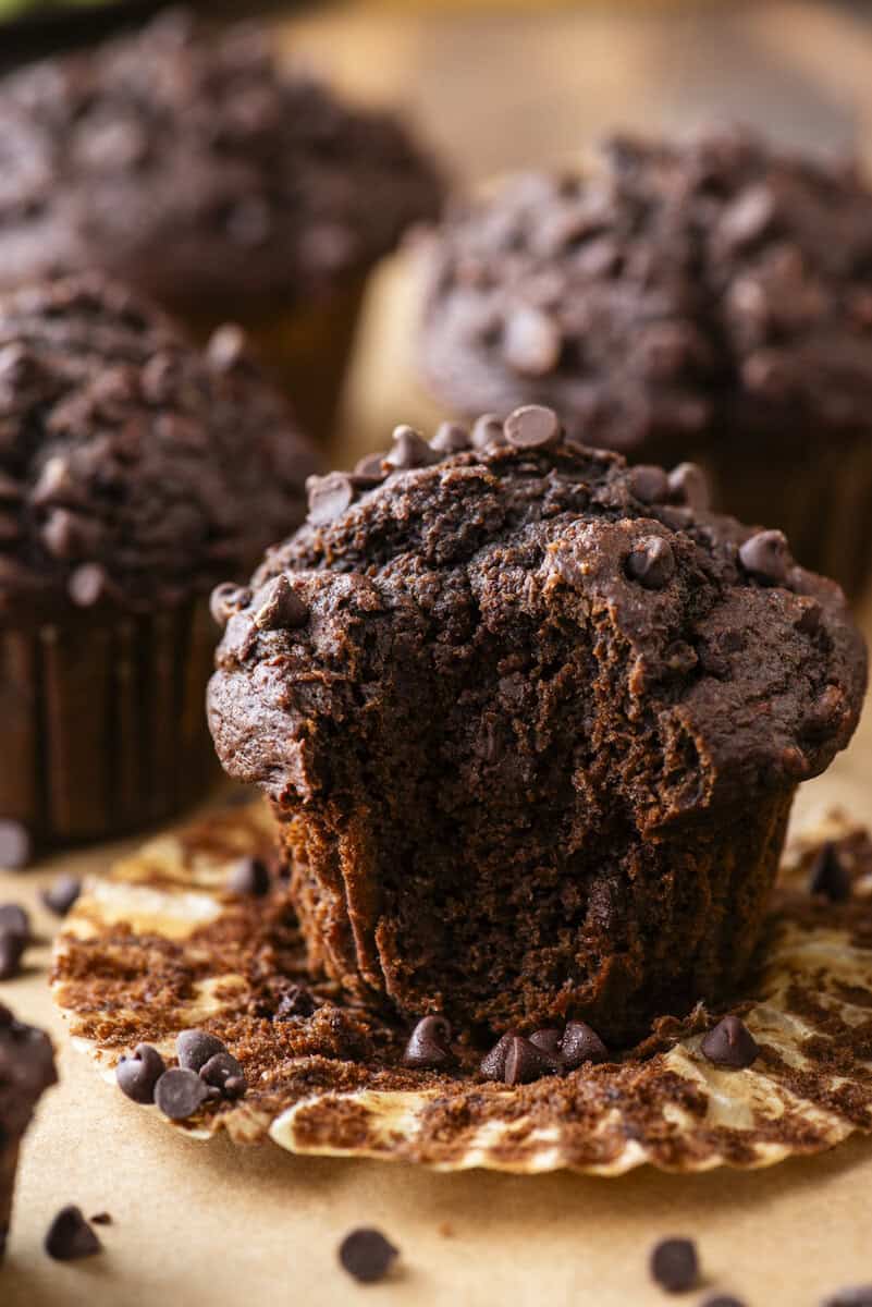 A close-up of a chocolate muffin with chocolate chips, partially eaten, sits on a crinkled muffin liner. More whole chocolate muffins are blurred in the background.