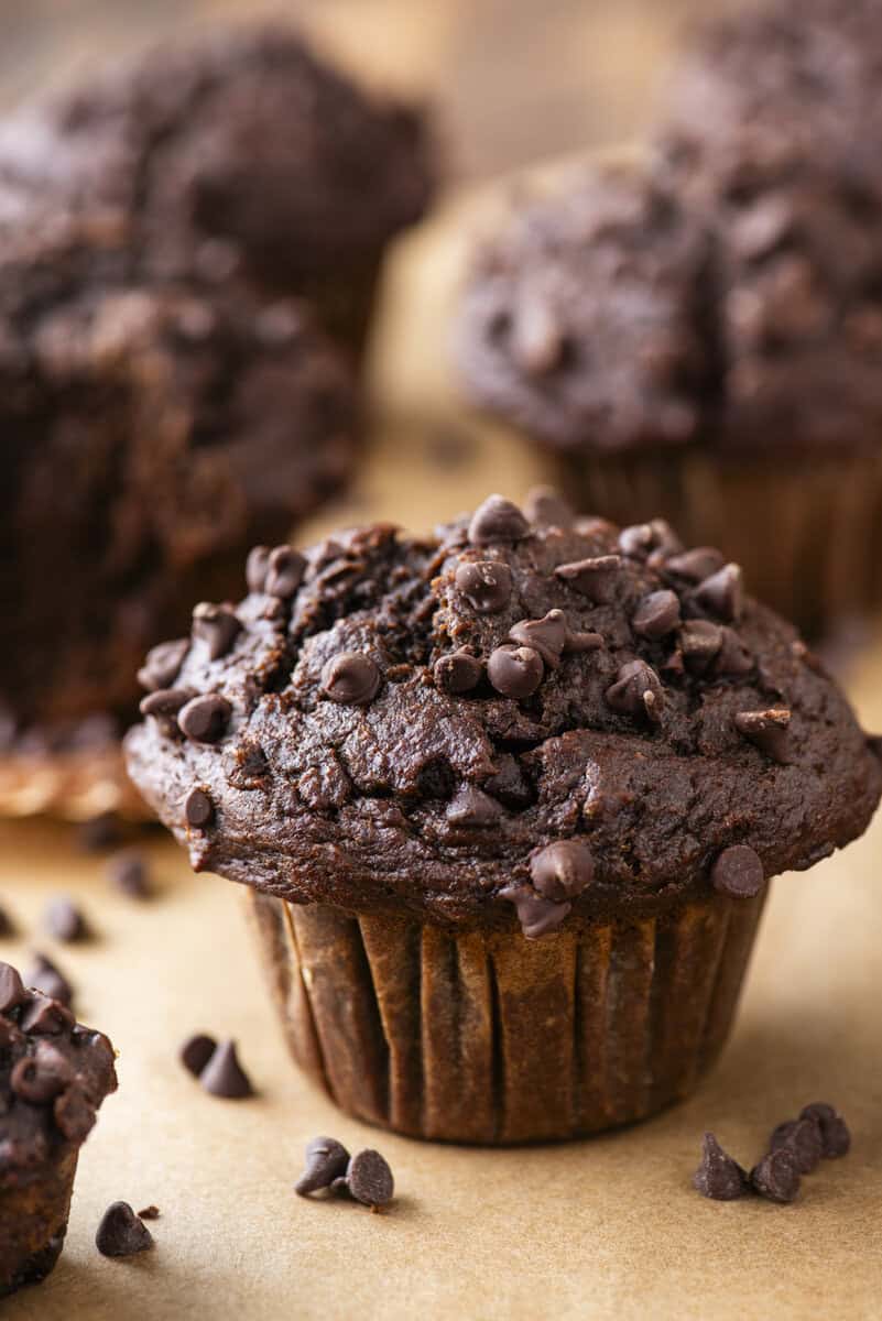 Chocolate muffin topped with small chocolate chips sits on brown parchment paper, with more muffins and scattered chocolate chips blurred in the background.