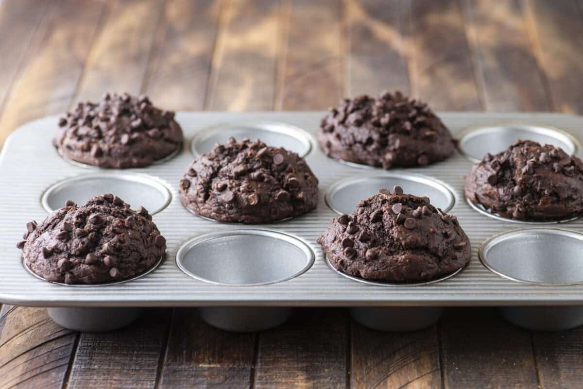 A metal muffin tray holds five chocolate muffins topped with chocolate chips, with three empty spaces. The tray sits on a rustic wooden surface.