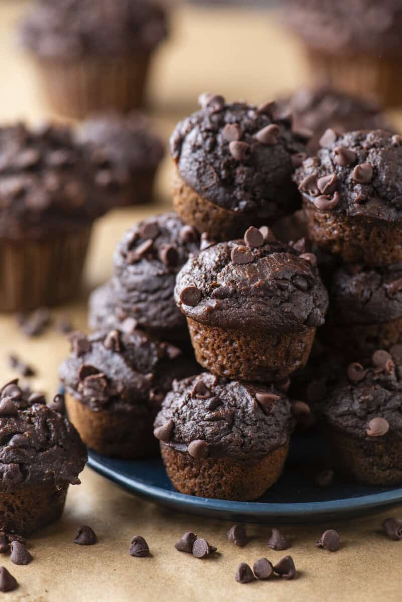 A plate stacked with mini chocolate muffins topped with chocolate chips, surrounded by more muffins on a brown surface.