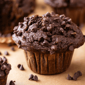 A close-up of a chocolate muffin topped with chocolate chips, sitting on parchment paper with more chocolate chips scattered around.