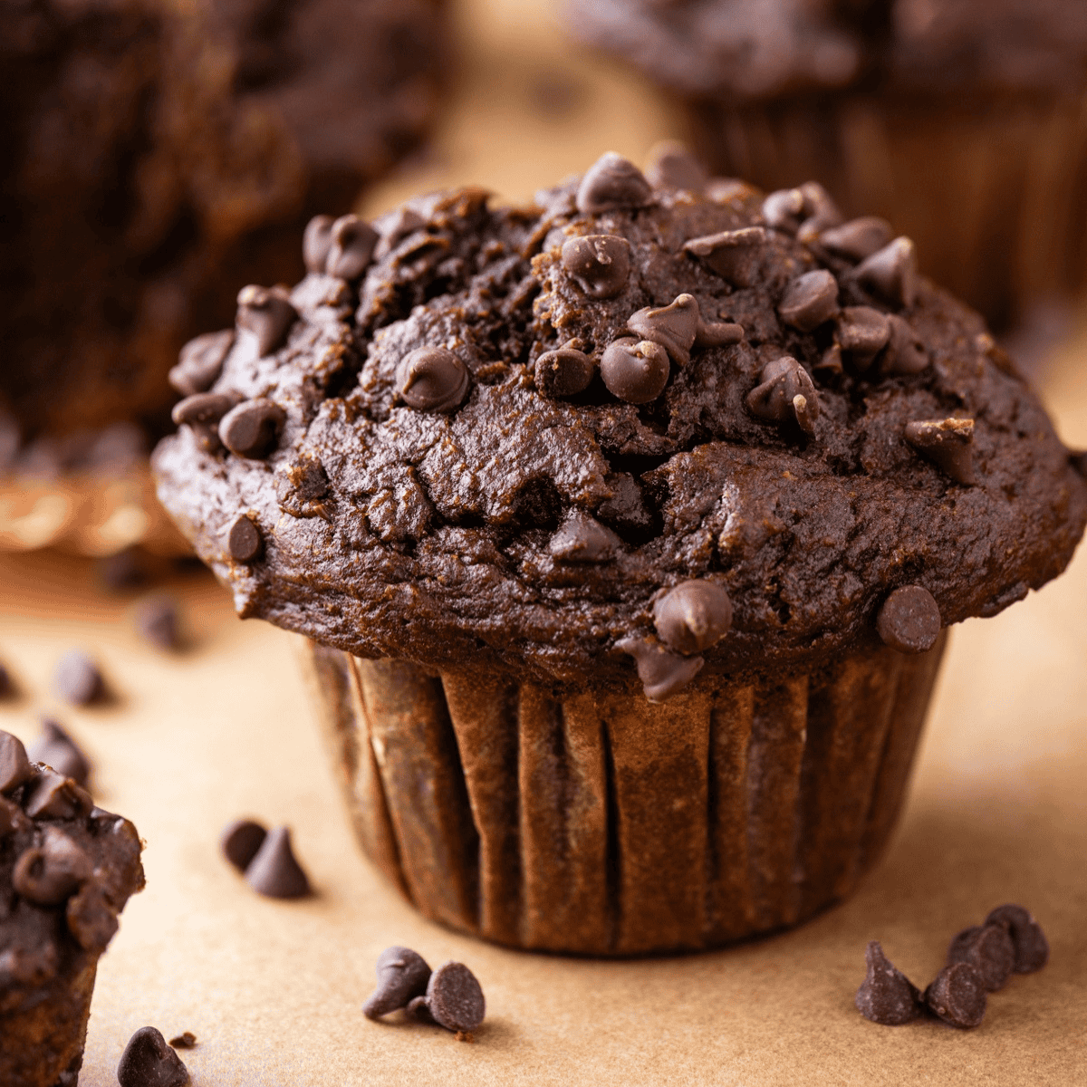 A close-up of a chocolate muffin topped with chocolate chips, sitting on parchment paper with more chocolate chips scattered around.