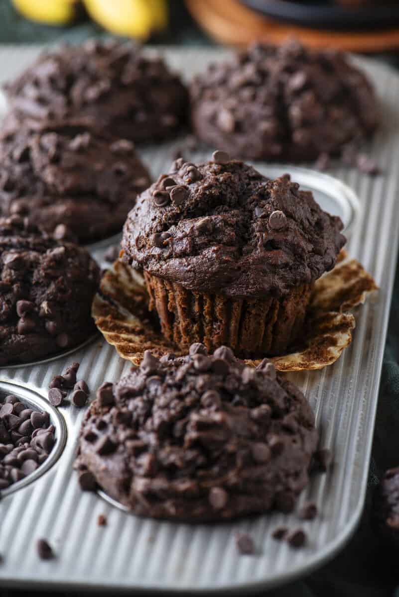 A close-up of chocolate muffins with chocolate chips in a muffin tin. One muffin is unwrapped, showing its moist texture.