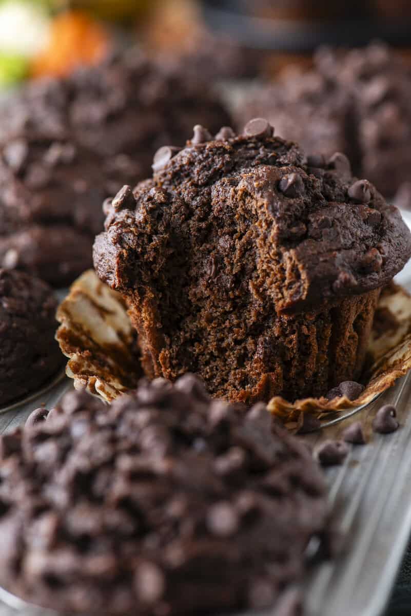 A close-up of a chocolate muffin with chocolate chips, partially unwrapped and missing a bite, surrounded by more chocolate muffins in the background.