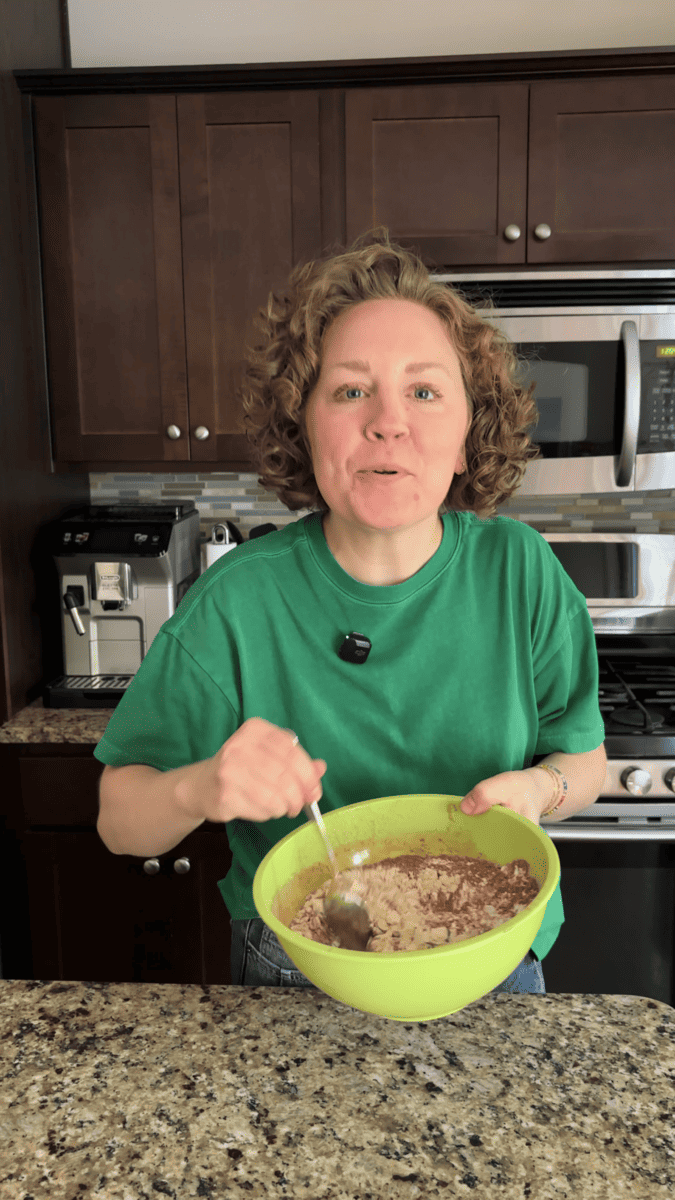 A woman in a green shirt stirs ingredients in a large green bowl while smiling in a kitchen with dark cabinets, a coffee maker, and a microwave visible in the background.
