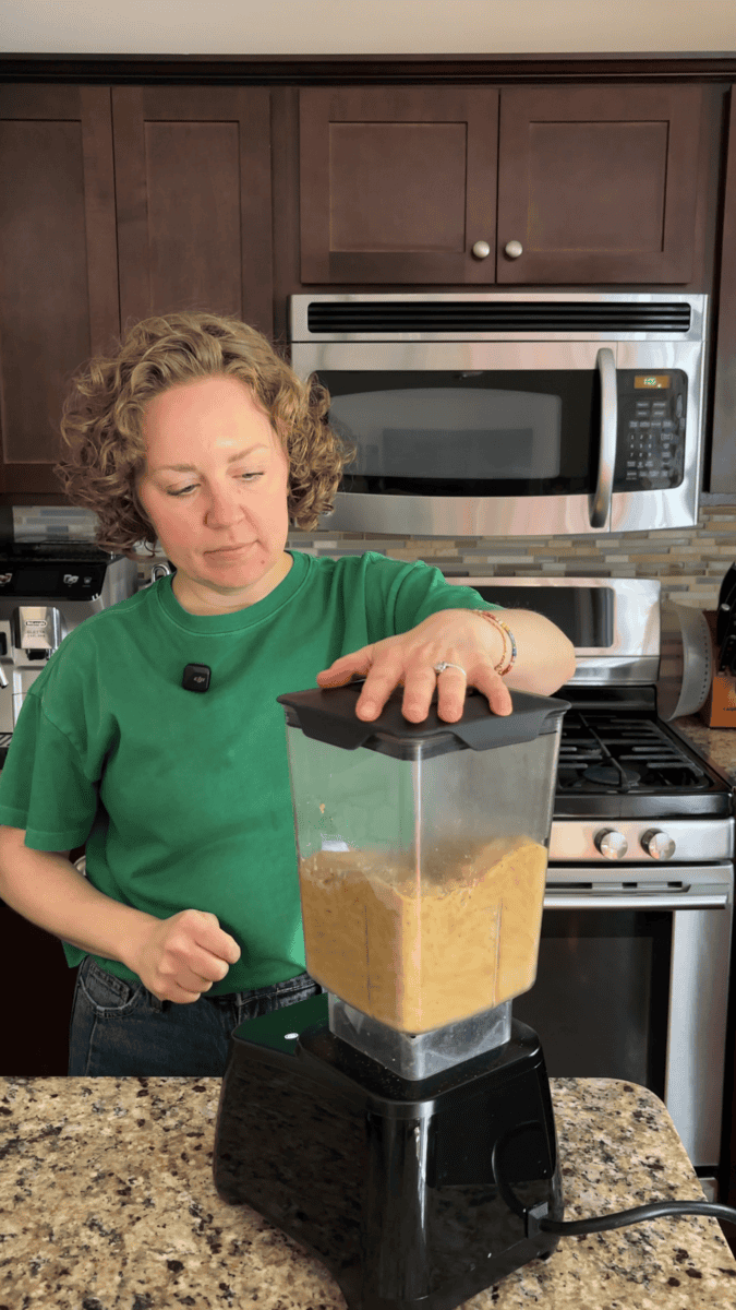 A person in a green shirt blends a mixture in a blender in a kitchen with brown cabinets and stainless steel appliances.