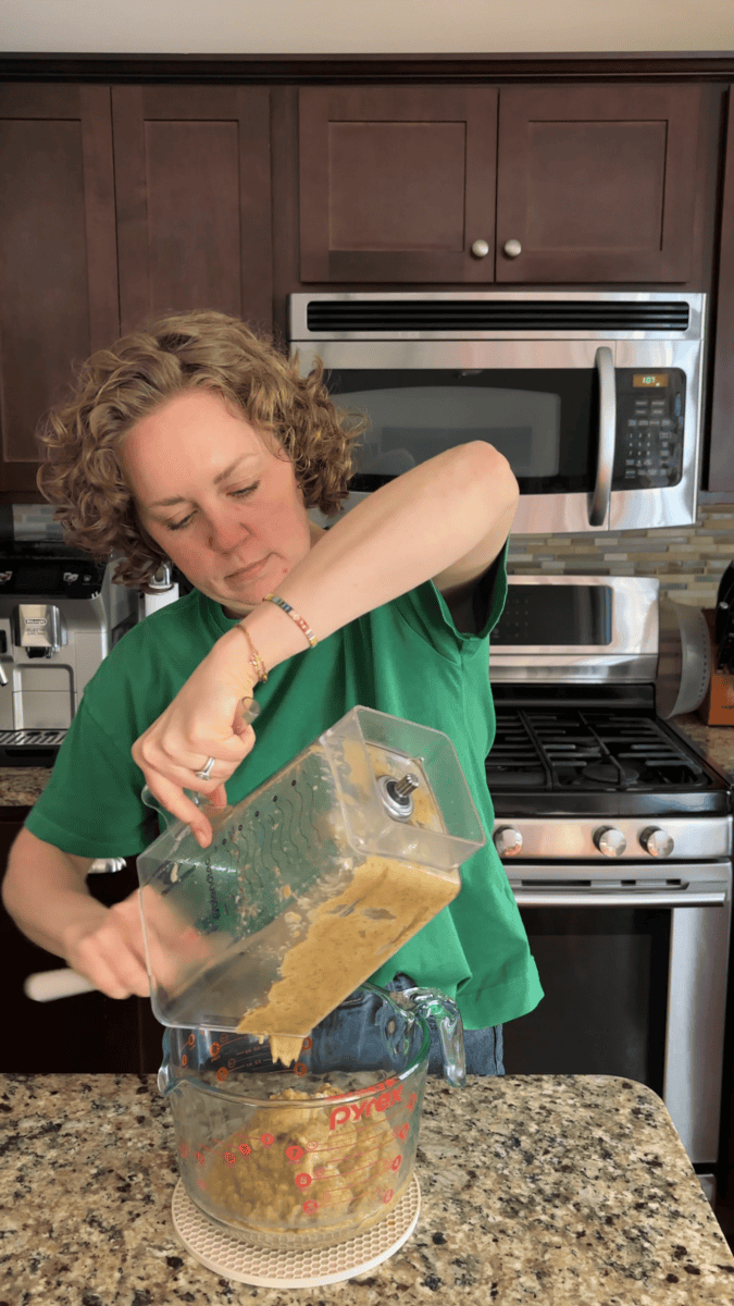 A person in a green shirt pours a thick blended mixture from a blender container into a glass measuring cup in a kitchen with dark cabinets and stainless steel appliances.