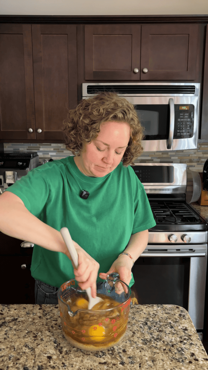 A person with curly hair wearing a green shirt stirs ingredients in a glass bowl in a kitchen, standing in front of a stove and microwave.