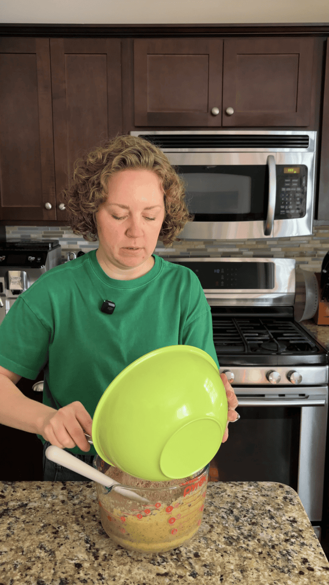 A person in a green shirt pours batter from a large green mixing bowl into a glass dish in a kitchen with dark cabinets and stainless steel appliances.