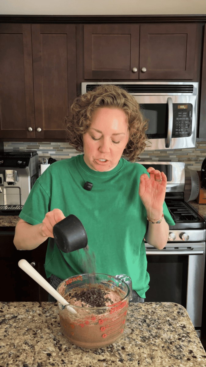 A person in a green shirt pours chocolate chips from a black measuring cup into a glass mixing bowl in a kitchen.