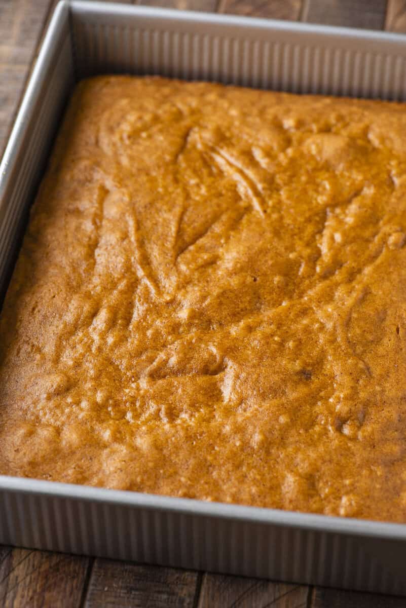 A square, golden-brown cake in a metal baking pan sits on a wooden surface. The cake has a slightly textured top with soft, even coloring and no frosting.