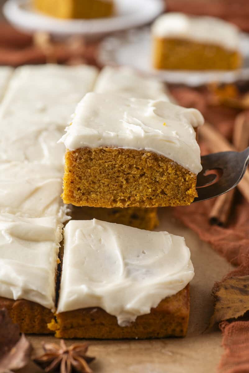 Pumpkin sheet cake with creamy white frosting sliced on parchment paper. A serving spatula lifts one square piece, showing the cakes moist texture. Cinnamon sticks and autumn leaves are in the background.
