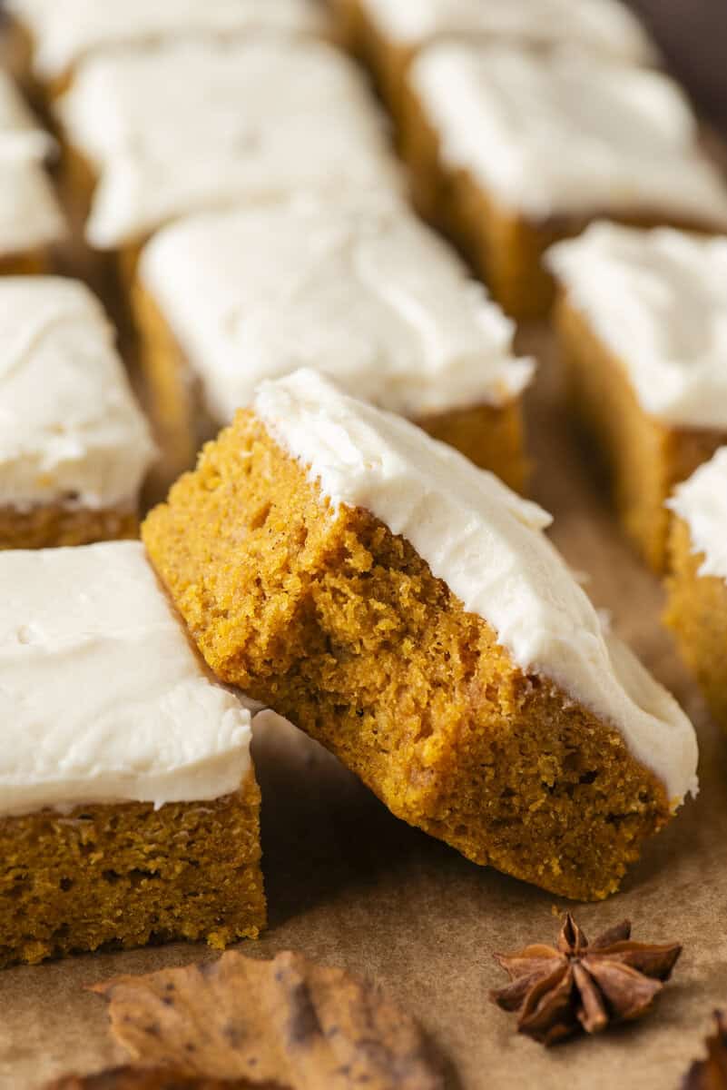 Pumpkin bars topped with creamy white frosting, arranged on brown parchment paper. One bar is propped up to show its moist texture. A star anise and a dry leaf sit nearby.