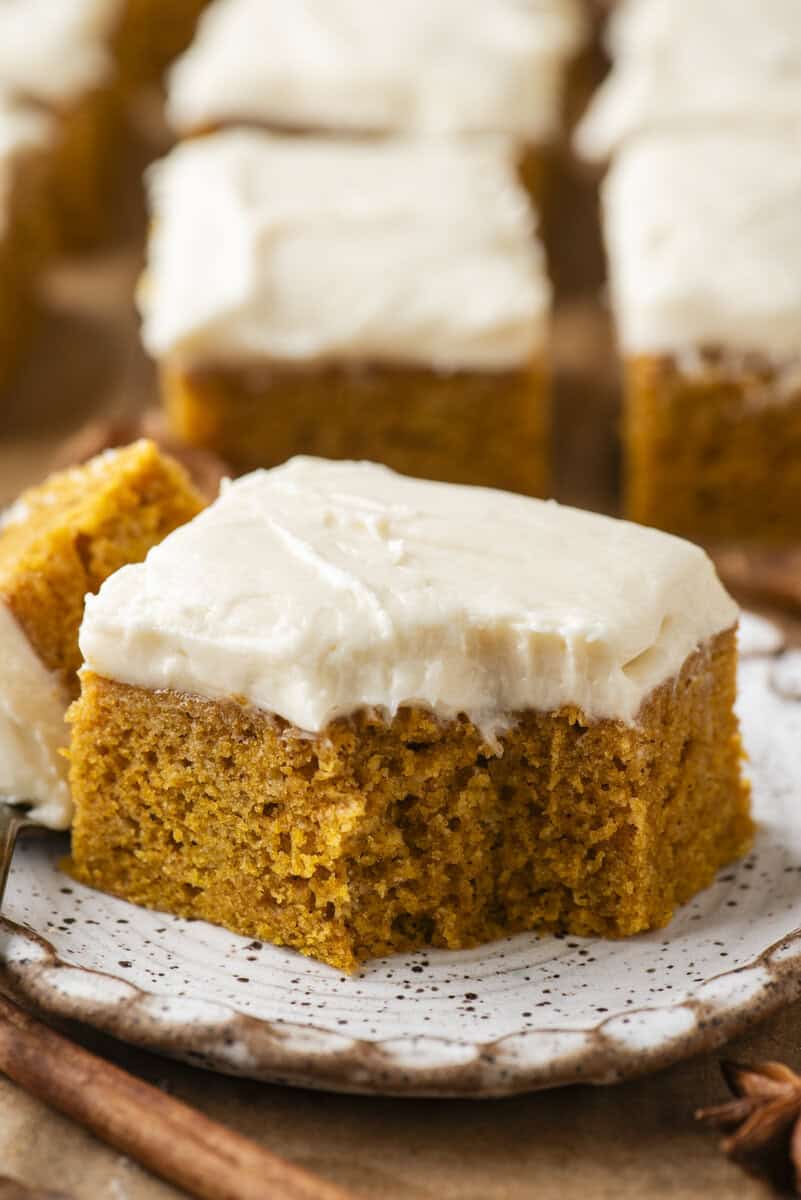 A square piece of pumpkin cake with creamy white frosting sits on a speckled plate. A bite has been taken from the cake, showing its moist texture. Other frosted cake pieces are in the background.