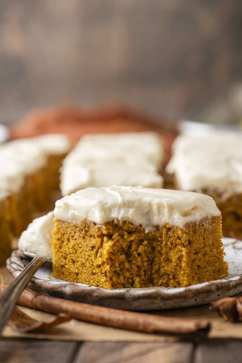 A close-up of a slice of pumpkin cake topped with creamy white frosting on a plate, with more frosted cake pieces and cinnamon sticks in the background.