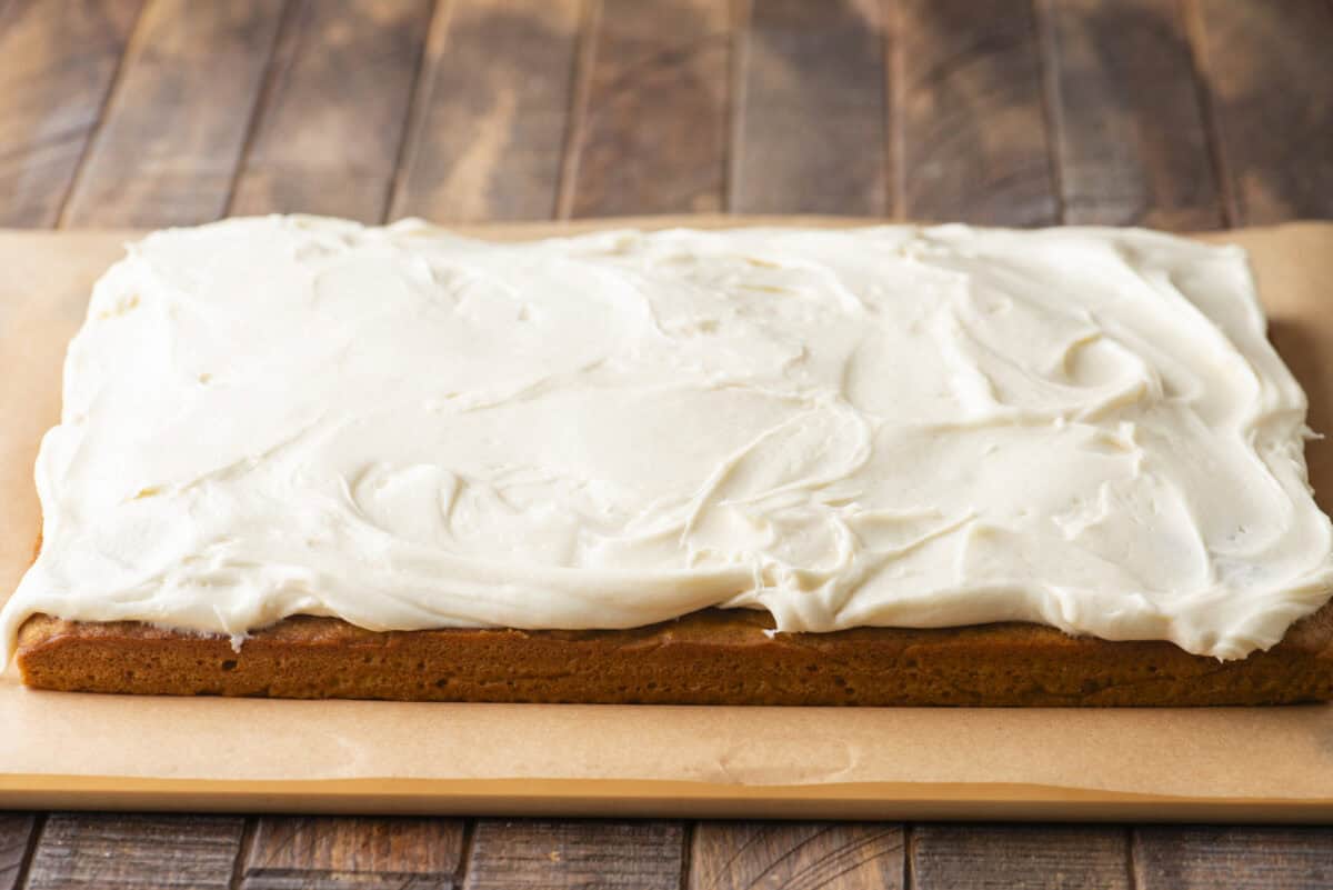 A rectangular sheet cake on parchment paper, topped with a thick, even layer of creamy white frosting, sits on a wooden surface.