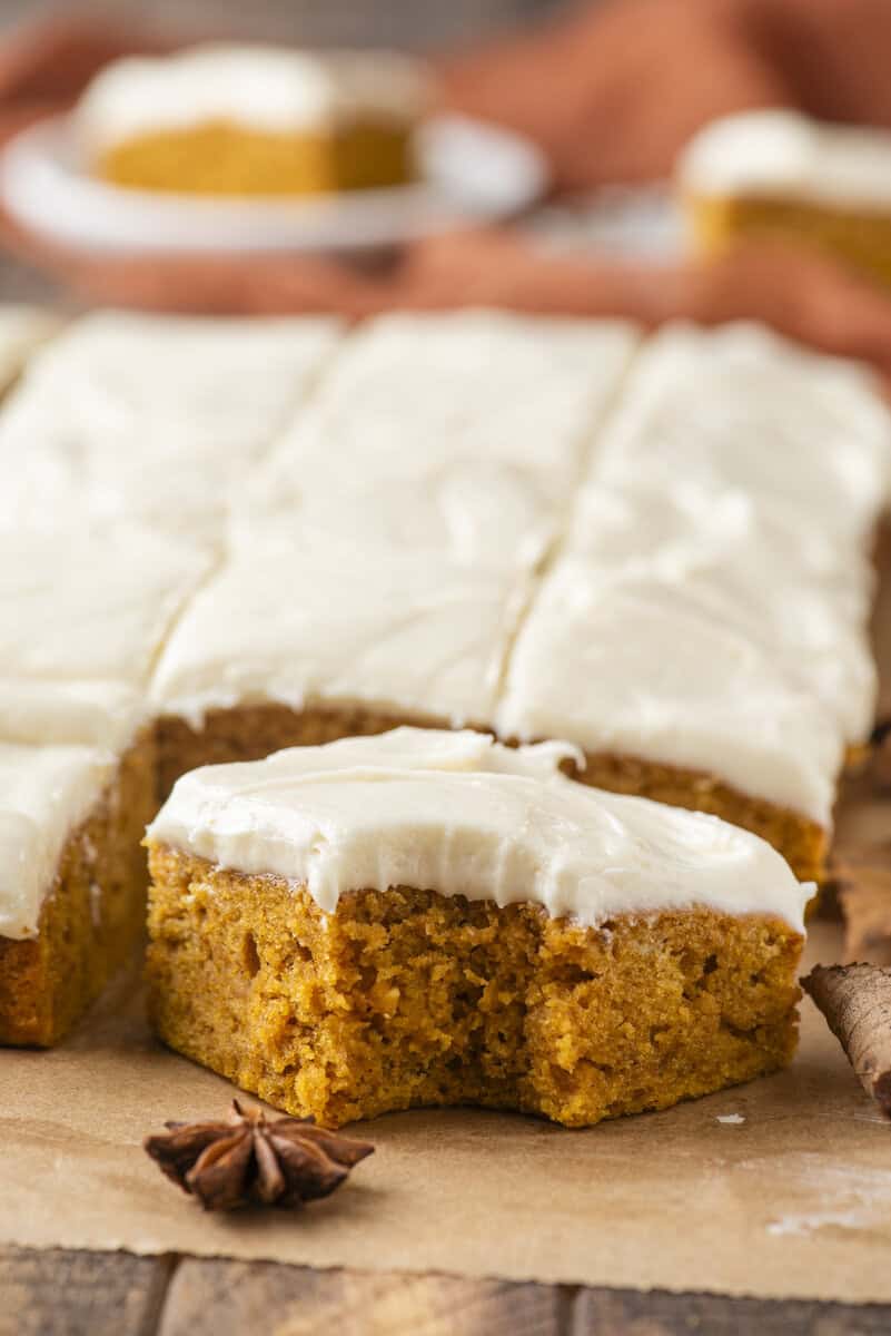 A close-up of pumpkin bars topped with creamy white frosting, with one bar removed to show its moist texture. A cinnamon stick and star anise are in the foreground.