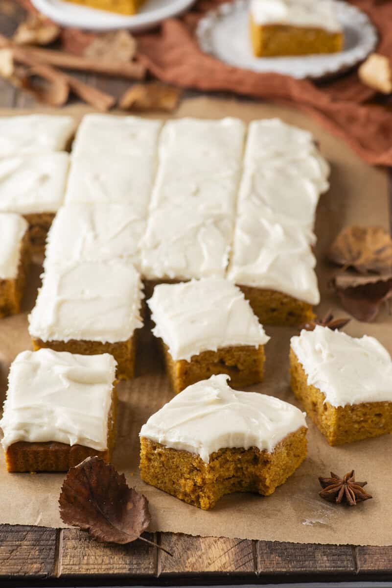 Pumpkin bars with white frosting cut into squares on parchment paper, surrounded by autumn leaves and spices. One bar in the foreground has a bite taken out of it.