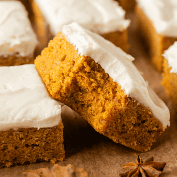 Close-up of pumpkin bars with creamy white frosting on top, arranged on brown parchment paper. One bar is leaning against another, showing its moist texture.