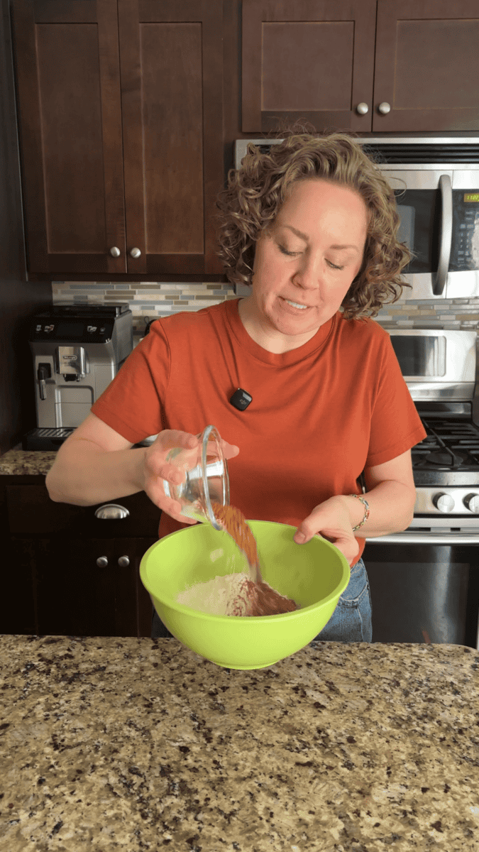 A woman in an orange shirt pours cocoa powder from a glass bowl into a green mixing bowl filled with flour in a kitchen with dark cabinets and stainless steel appliances.
