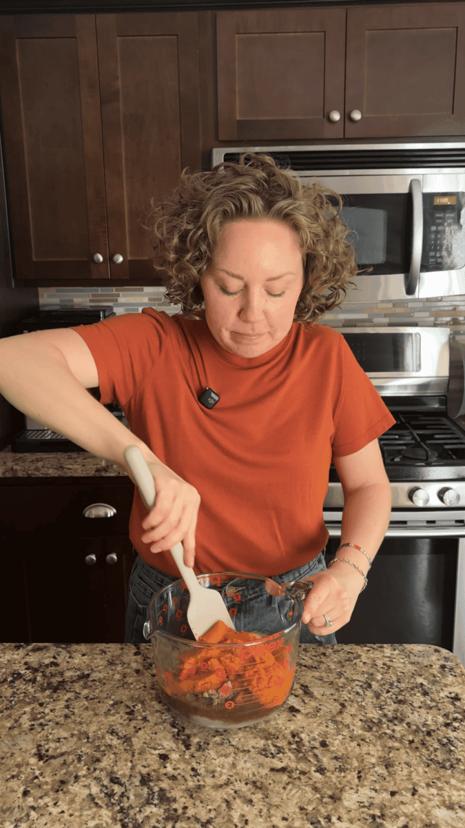 A woman in an orange shirt stirs ingredients in a glass bowl with a spatula in a kitchen, standing at a countertop with a stove and microwave behind her.