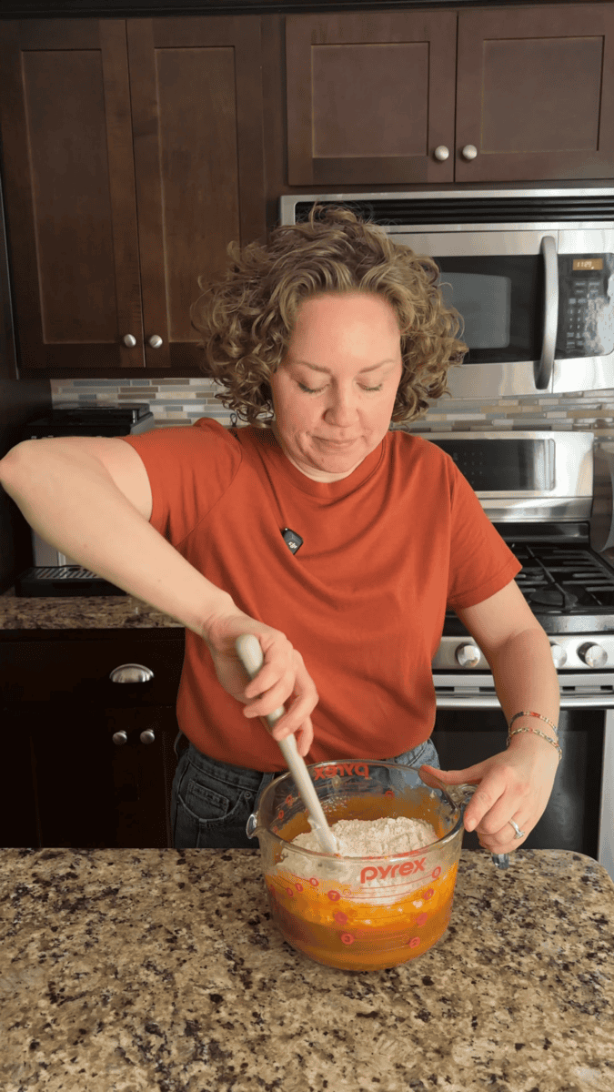 A person with curly hair in an orange shirt mixes ingredients in a large glass measuring cup in a kitchen with dark cabinets and a stainless steel stove.
