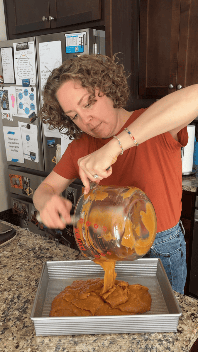 A woman pours thick batter from a glass bowl into a metal baking pan in a kitchen, standing by a granite countertop with a fridge and cabinets in the background.